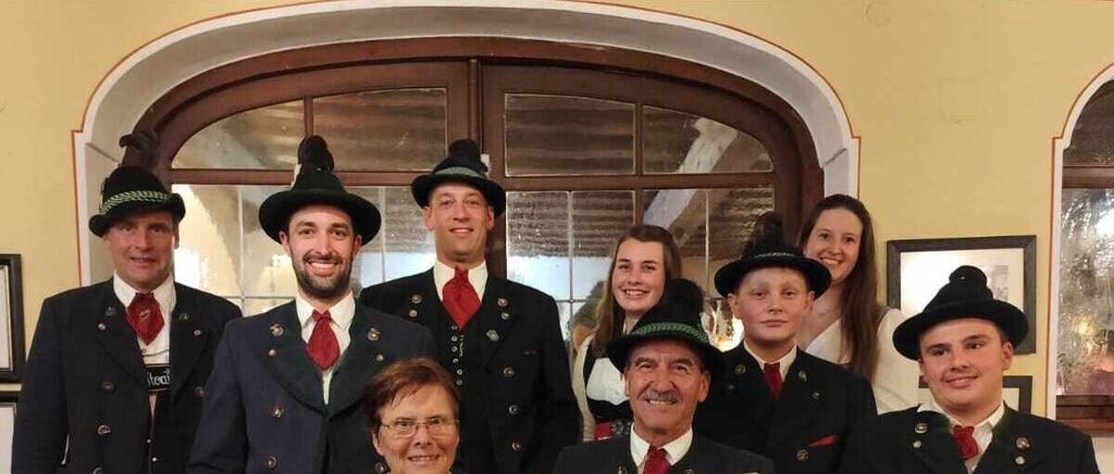 A group of people dressed in traditional German attire poses for a photo in front of a brown door. Some wear hats, glasses, and red ties. A smiling woman stands in the center.