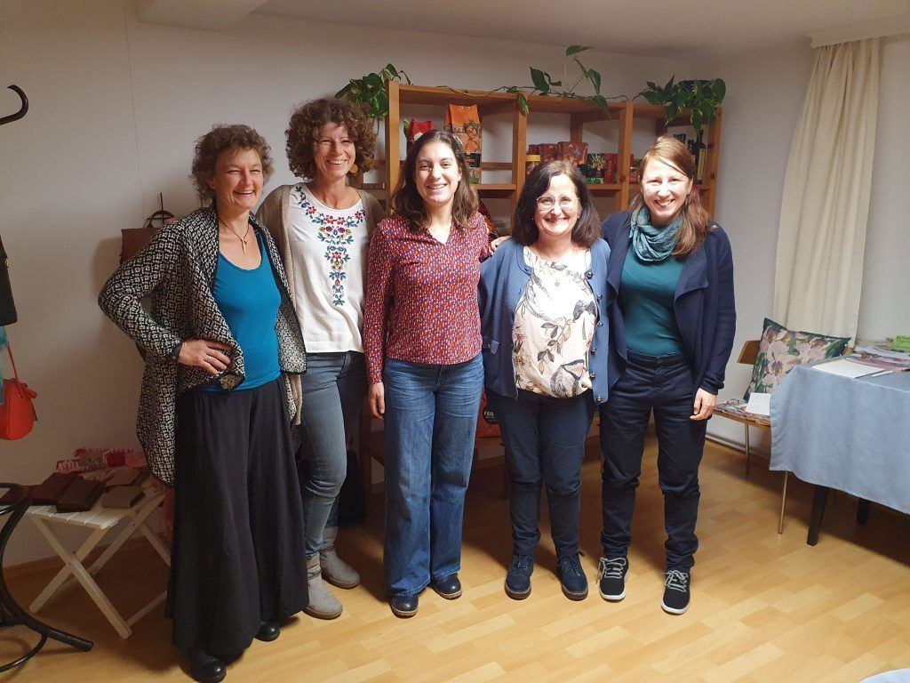 Five women stand together in a room, smiling for a photo. Behind them is a wooden shelf with plants and books. The floor is wooden.