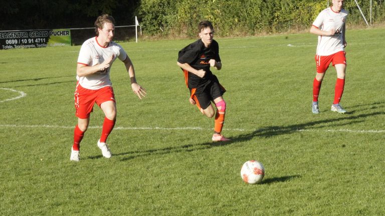 Zwei männliche Fußballspieler auf einem Feld, einer jagt dem Ball nach. Der Spieler in Schwarz läuft mit dem Ball, während der Spieler in Weiß ihn verfolgt.
