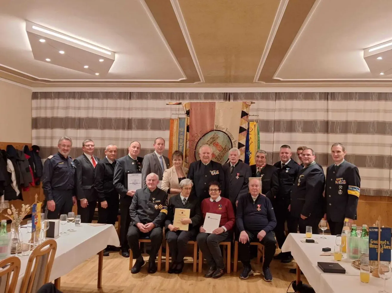 A group of formally dressed individuals stand and sit in a banquet hall, with some holding certificates and glasses of wine. Flags hang on the wall behind them.