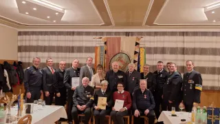 A group of formally dressed individuals stand and sit in a banquet hall, with some holding certificates and glasses of wine. Flags hang on the wall behind them.