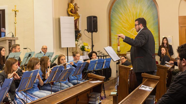 Ein Dirigent steht vor einer Gruppe junger Flötenspieler in einer Kirchenumgebung. Hinter ihm sind ein Lautsprecher und eine Kerze auf einem Ständer zu sehen.