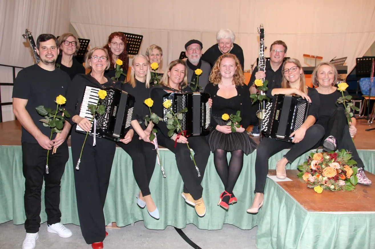 A group of musicians with black outfits and glasses are smiling and posing for a photo with their instruments, including an accordion and a clarinet. They are sitting on a bench covered with a green cloth.