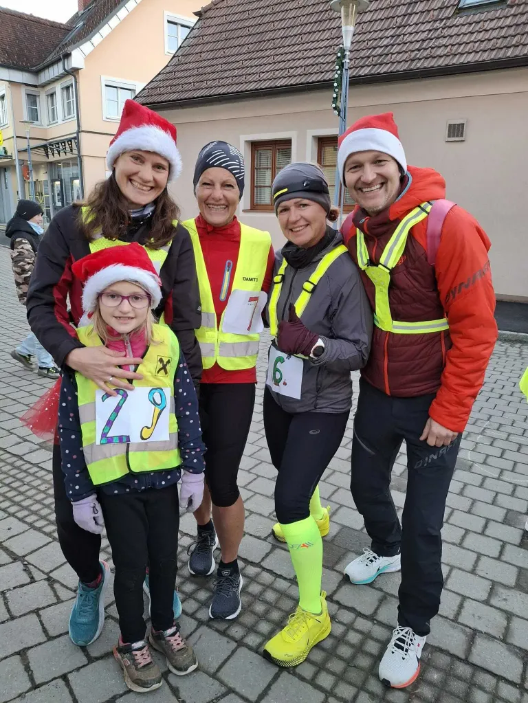Fünf Menschen in Weihnachtsmützen und Laufausrüstung stehen auf einem gepflasterten Weg. Sie lächeln und scheinen für ein Foto zu posieren. Das Mädchen in der Mitte hat die Nummer 29 auf ihrer Weste.