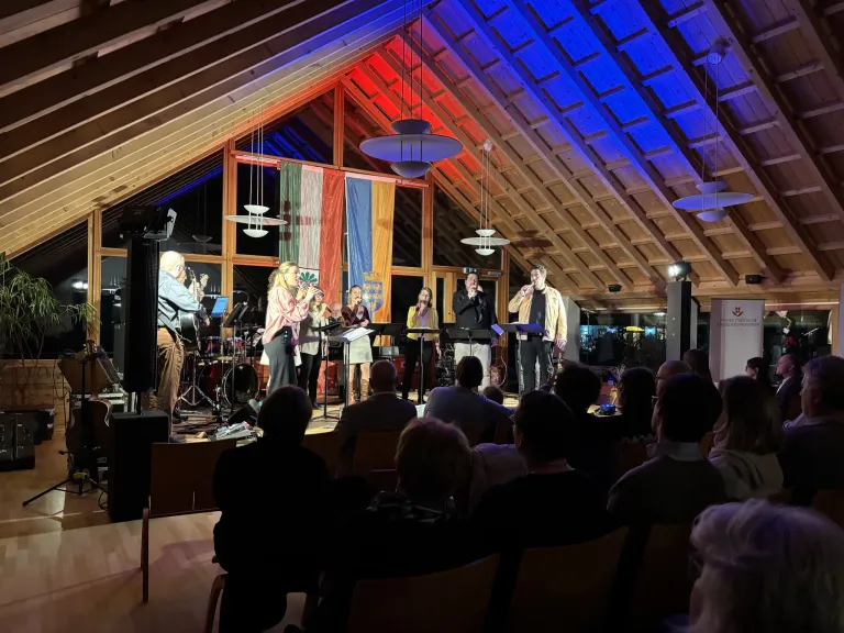 A band is performing on a stage with an audience in an attic room with wooden beams and colorful lights.