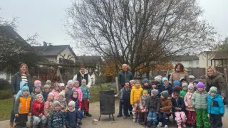 A group of children and two adults are gathered in front of a house, posing for a photo. They are all dressed warmly in winter clothing. Behind them is a tree and a trash can.