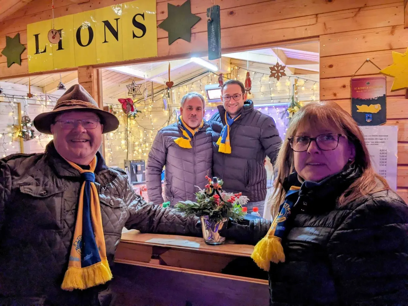 Three people in winter clothes pose in front of a decorated booth, with two men wearing yellow and blue scarves and one woman wearing glasses. The booth has a yellow sign with the word 'IONS' and a star. The booth is decorated with Christmas lights and ornaments.