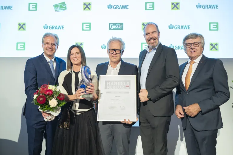 Eine Gruppe von fünf Personen posiert für ein Foto mit Auszeichnungen und einem Zertifikat in der Hand. Eine Frau in der Mitte hält eine Trophäe. Hinter ihnen zeigt eine Kulisse Logos und Text.