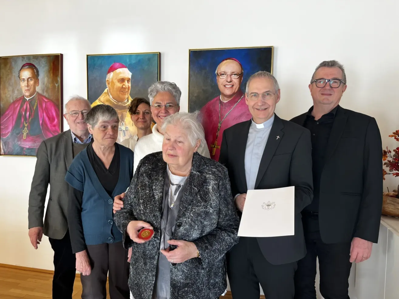 Eine Gruppe von Personen, darunter Geistliche, steht vor Porträts religiöser Persönlichkeiten. Eine ältere Frau hält ein Zertifikat und eine Medaille.
