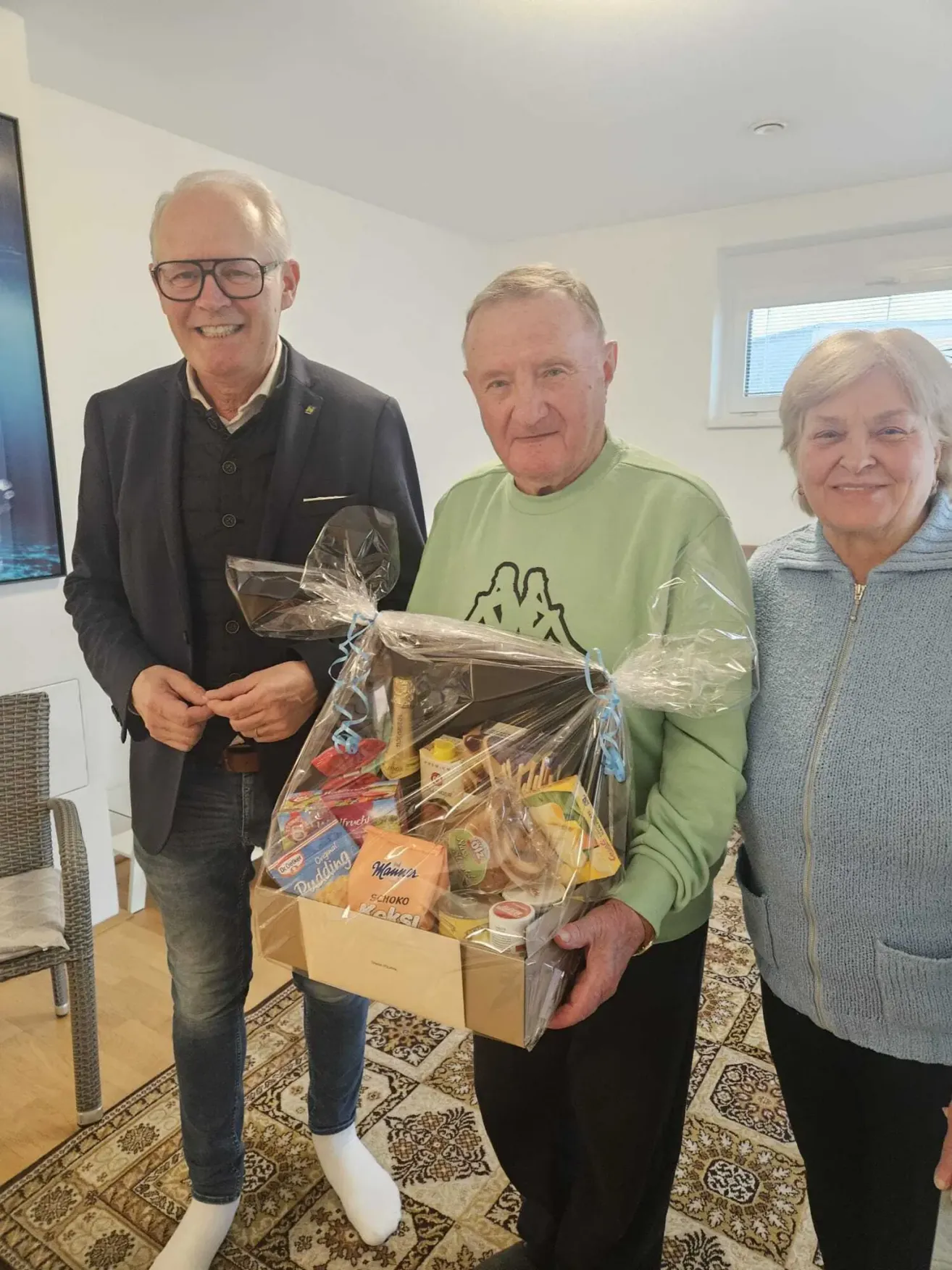 Drei Personen stehen in einem Raum mit einem Geschenkkorb. Ein Mann auf der linken Seite trägt eine Brille und einen Blazer, der Mann in der Mitte hält einen Korb voller Snacks und eine Frau auf der rechten Seite lächelt. Hinter ihnen ist eine Wand mit einem Gemälde und einem Fenster.