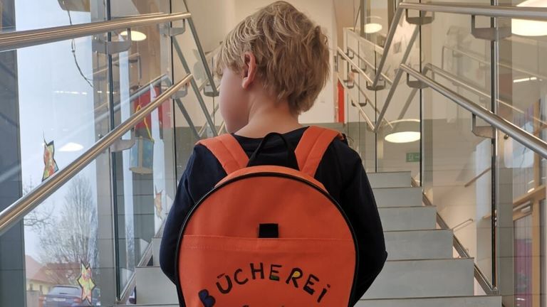Ein Kind mit einem Rucksack, auf dem Bucherei steht, geht eine Treppe mit einer Glasbrüstung hinunter.