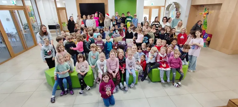 Eine große Gruppe von Kindern in Winterkleidung posiert für ein Foto, einige sitzen auf grünen Bänken, während andere stehen und Geschenke halten, in einem Raum mit grünen Wänden und Erwachsenen in der Nähe.