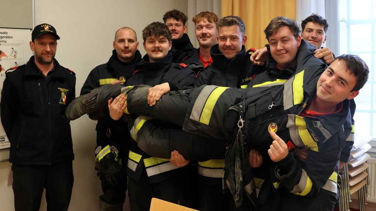 Eine Gruppe von Feuerwehrleuten posiert für ein Foto mit einer Person, die von anderen getragen wird, in einem Raum.