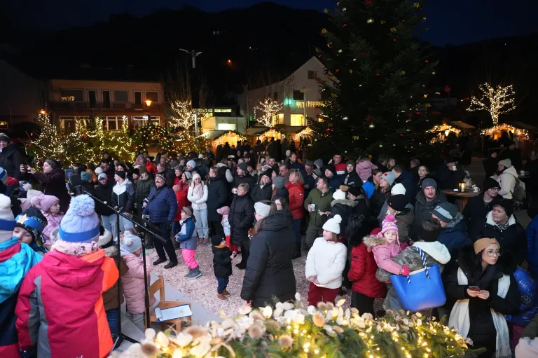 Eine Menschenmenge in Winterkleidung versammelt sich bei Nacht um einen Weihnachtsbaum. Das Gebiet ist mit festlichen Lichtern und Bäumen geschmückt. Einige Leute halten Tassen. Es gibt ein Mikrofon auf einem Ständer.
