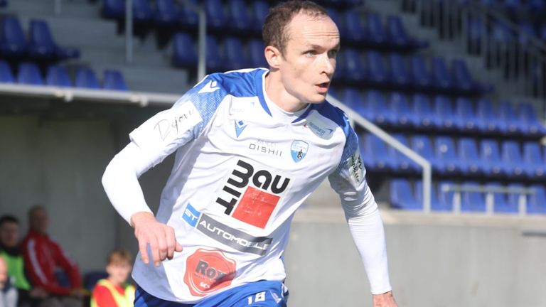 Ein männlicher Fußballspieler läuft auf dem Feld. Er trägt ein weißes Trikot mit verschiedenen Logos und Nummern. Der Hintergrund ist ein Stadion mit leeren blauen Sitzen.