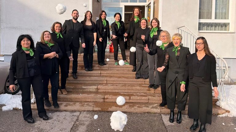 Eine Gruppe von Menschen in formeller Kleidung, die grüne Schals tragen, steht auf Stufen mit schneeklumpenartigen Baumwollbällen, die um sie herum schweben. Dahinter ist ein Gebäude mit einer Glastür zu sehen.