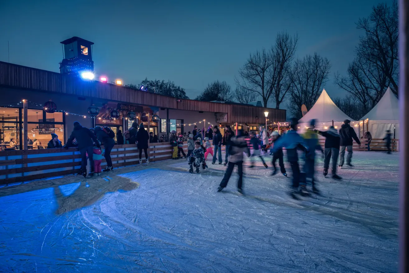 Bei Nacht sind mehrere Menschen auf einer Eisbahn im Freien beim Eislaufen. Es gibt ein Gebäude mit dem Wort 'SUNBAR' und einige dekorative Lichter. Ein weißes Zelt befindet sich in der Nähe.