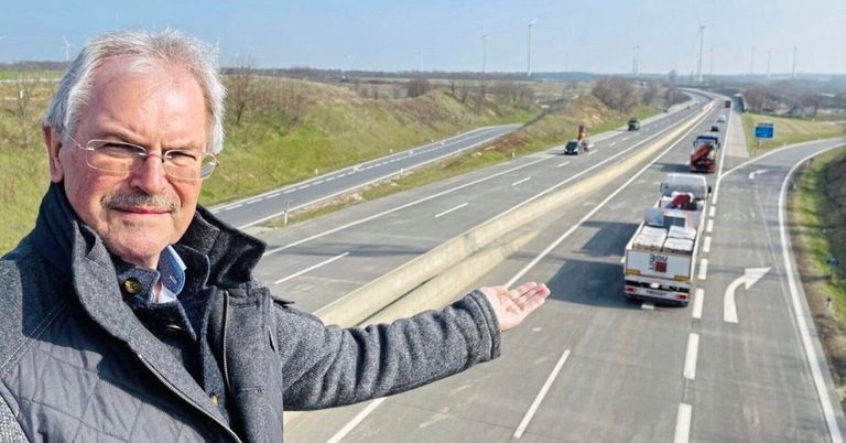 Ein älterer Mann mit Brille steht auf einer Brücke und zeigt mit der Hand auf eine Autobahn mit zwei Spuren, auf der Autos und Lastwagen fahren. In der Ferne sind Windturbinen zu sehen.