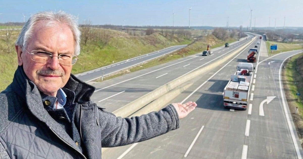 Ein älterer Mann mit Brille steht auf einer Brücke und zeigt mit der Hand auf eine Autobahn mit zwei Spuren, auf der Autos und Lastwagen fahren. In der Ferne sind Windturbinen zu sehen.