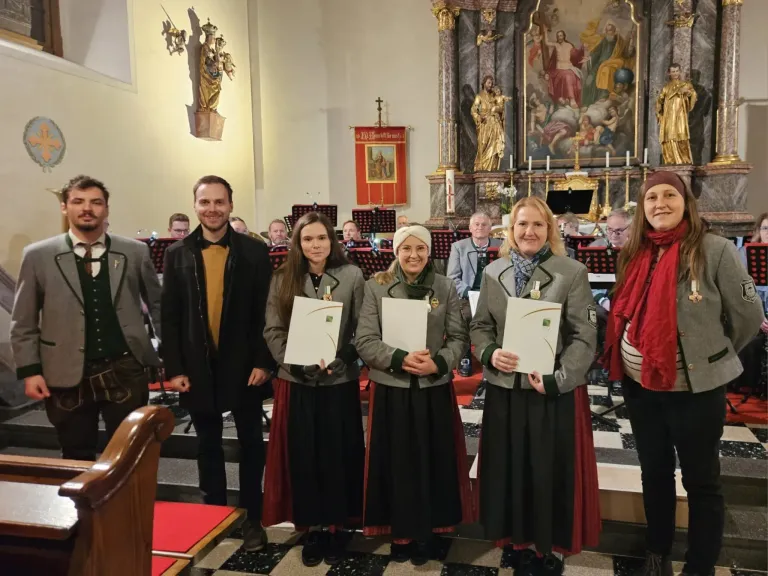 Drei Frauen und ein Mann stehen auf einem gemusterten Boden und halten Urkunden in den Händen. Sie lächeln und stehen vor einer Kircheninnenansicht. Hinter ihnen sitzen Leute auf Stühlen und ein großes Gemälde an der Wand.
