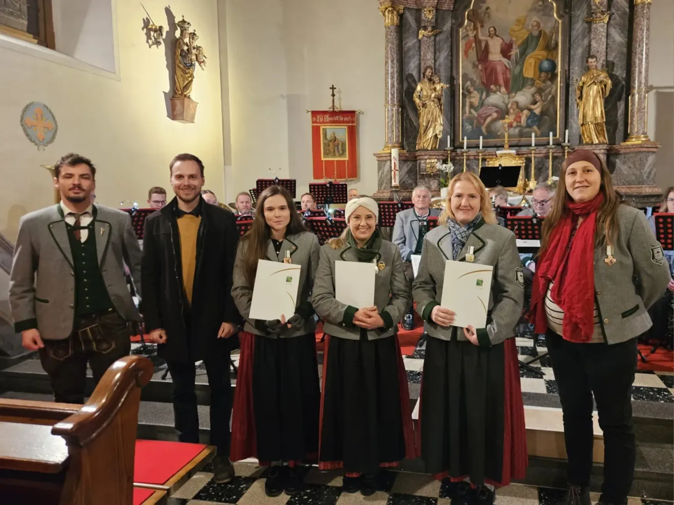 Drei Frauen und ein Mann stehen auf einem gemusterten Boden und halten Urkunden in den Händen. Sie lächeln und stehen vor einer Kircheninnenansicht. Hinter ihnen sitzen Leute auf Stühlen und ein großes Gemälde an der Wand.