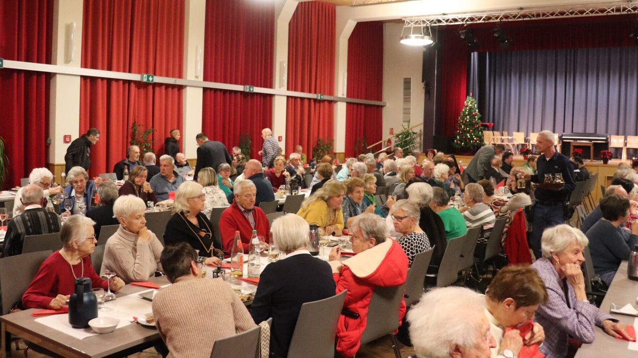 Eine Gruppe älterer Menschen versammelt sich in einem Speisesaal, sitzt an Tischen und führt Gespräche. Auf der Bühne ist ein Weihnachtsbaum zu sehen.