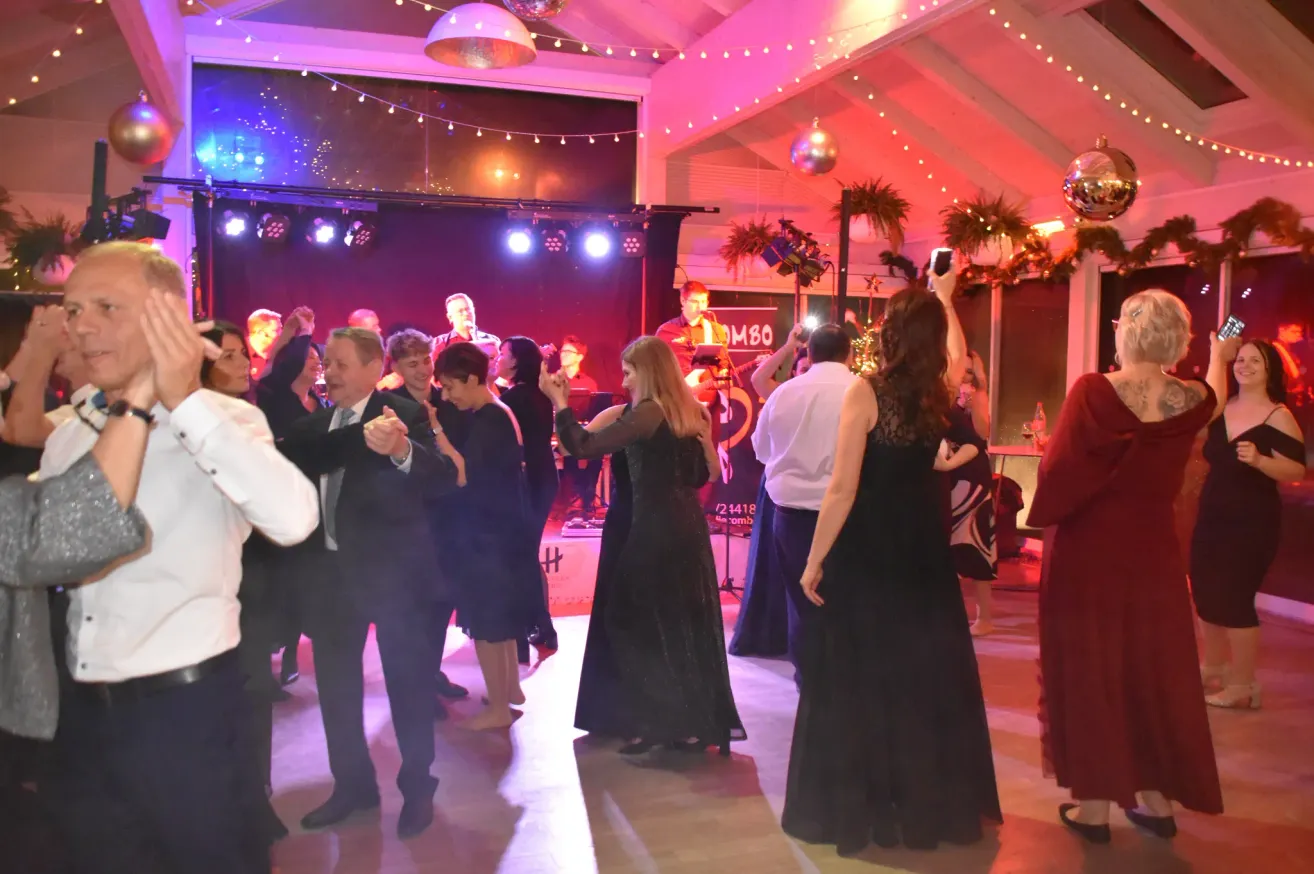 Menschen tanzen auf einer Party mit Bühne, Disko-Lichtern und festlicher Atmosphäre. Eine Frau in Schwarz führt den Tanz an, während andere um sie herum mittanzen.