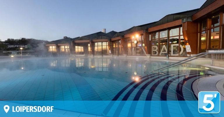 Ein Außenpoolbereich im Menbad-Komplex, umgeben von großen Glasfenstern, mit einer blauen Wasseroberfläche, Reflexionen und einem modernen Gebäudedesign.