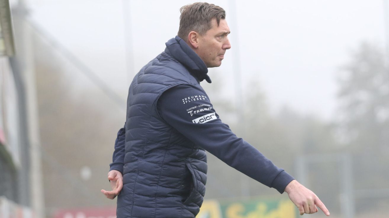 Ein Mann in einer dunkelblauen Jacke mit 'Reummann Fliesen' und 'GASTRO' auf dem linken Arm scheint jemanden auf einem nebligen Feld zu instruieren.