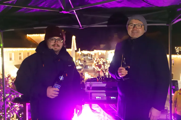 Zwei Männer stehen unter einem lila Zelt, lächeln und halten Flaschen. Hinter ihnen befindet sich ein DJ-Pult mit einem lila Licht. Der Bereich ist mit Weihnachtslichtern geschmückt und es sind Gebäude in der Ferne zu sehen.