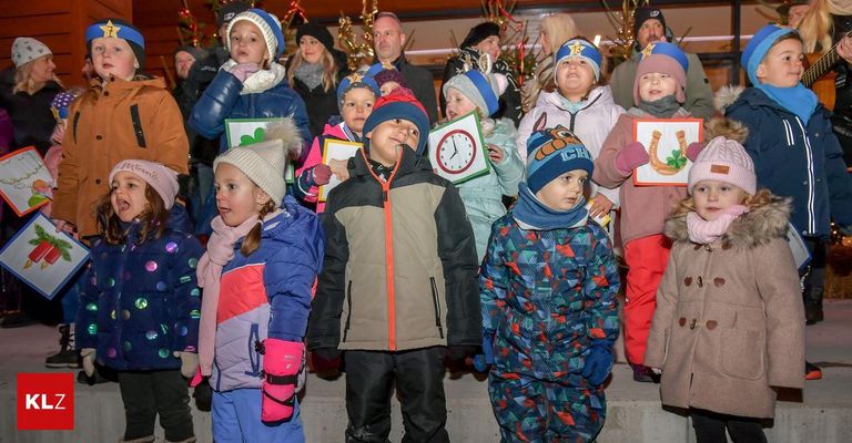 Eine Gruppe von Kindern und Erwachsenen steht zusammen und trägt Winterkleidung. Sie halten Karten hoch. Einige der Kinder tragen Mützen, und sie stehen vor einem Gebäude mit Weihnachtsdekoration.