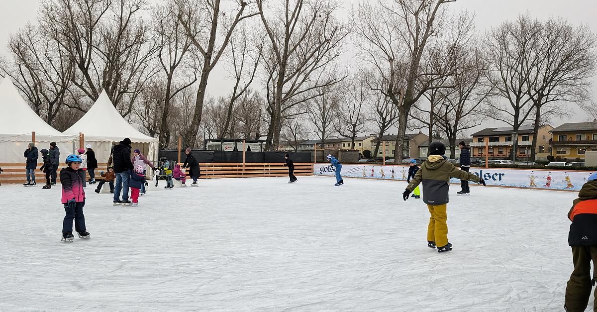 Mehrere Menschen eislaufen auf einer Freiluftbahn, umgeben von kahlen Bäumen und Gebäuden. Einige tragen Mützen und Handschuhe. Ein Zaun begrenzt die Bahn.