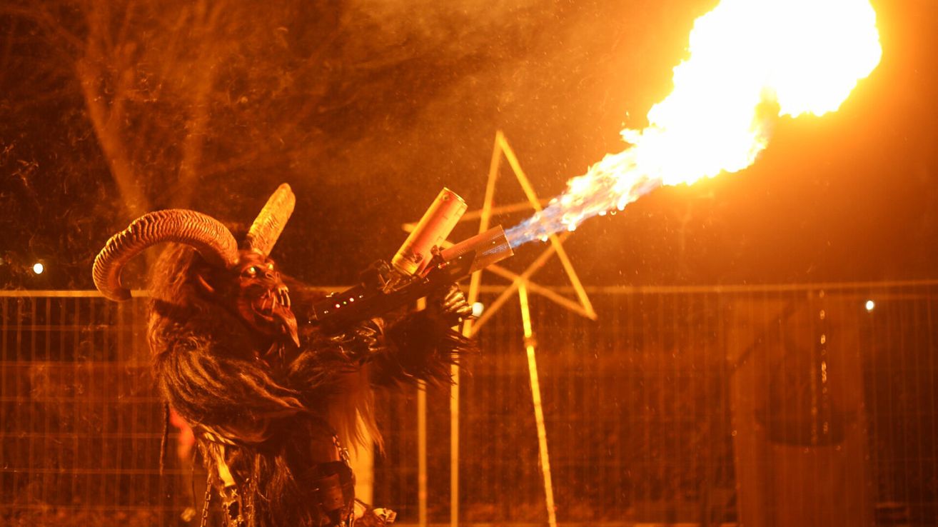 Eine Person in einem gehörnten Kostüm, die einen Flammenwerfer hält und Feuer in die Luft schießt, mit einem sternförmigen Muster hinter ihr.