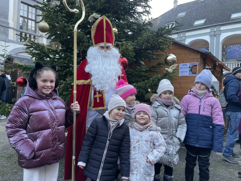 Eine Gruppe von Kindern und ein als Weihnachtsmann verkleideter Mann stehen vor einem Weihnachtsbaum. Die Kinder tragen Winterkleidung. Hinter ihnen befindet sich ein hölzerner Stand mit einem Schild, auf dem "Guesch Hotte" steht. Im Hintergrund befindet sich ein Gebäude.