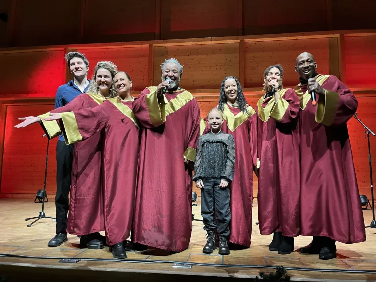 Ein Chor in roten und goldfarbenen Roben steht auf der Bühne, mit einem jungen Jungen in Grau in der Mitte. Dahinter sorgen eine Holzwand und rote Beleuchtung für eine festliche Atmosphäre.