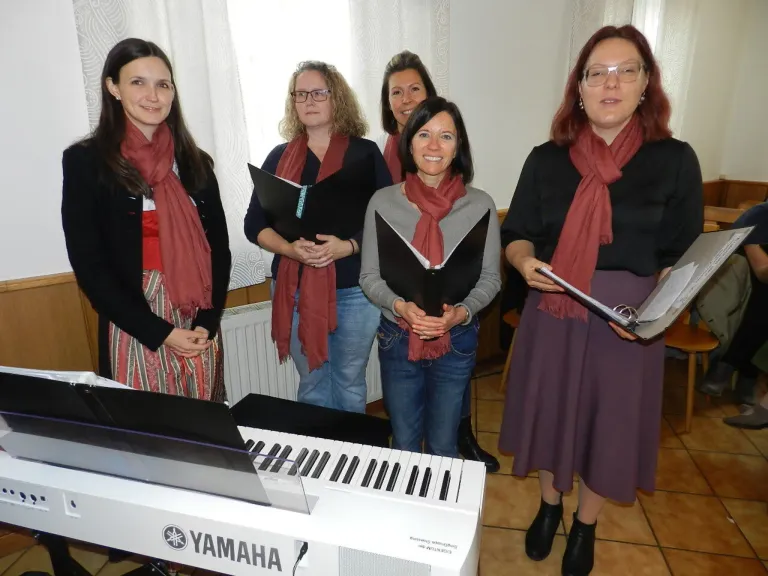 Fünf Frauen stehen hinter einem Yamaha-Piano, tragen Schals und halten Notenblätter in der Hand, lächeln für ein Foto in einem Raum mit weißen Vorhängen.