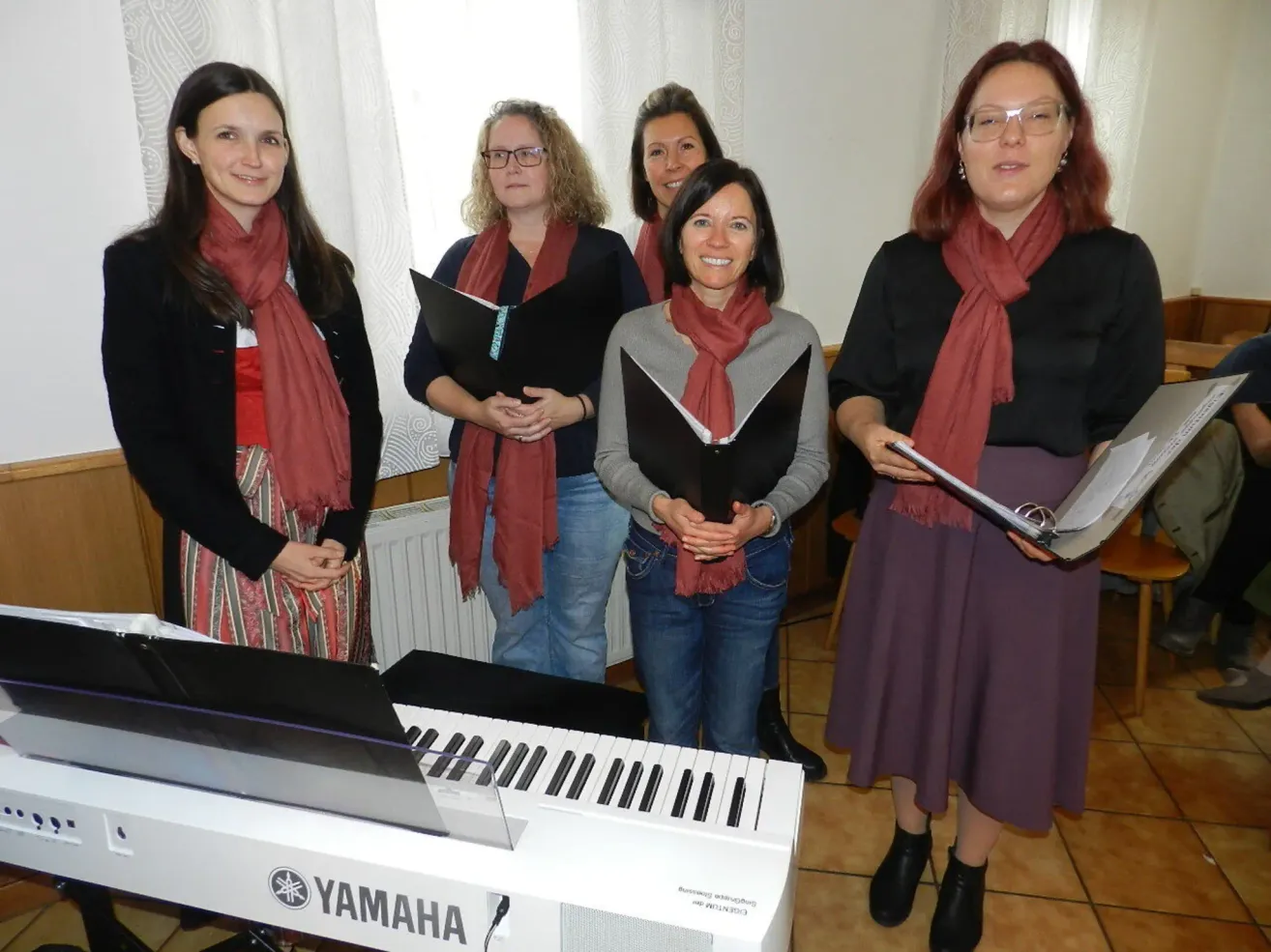 Fünf Frauen stehen hinter einem Yamaha-Piano, tragen Schals und halten Notenblätter in der Hand, lächeln für ein Foto in einem Raum mit weißen Vorhängen.