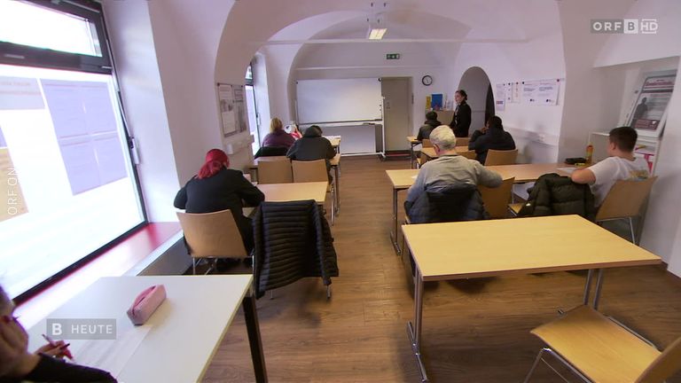 Ein Klassenzimmer mit gewölbten Decken, Schülern in Reihen und einer Lehrerin, die an einer Tafel steht. An den Wänden hängen Zettel.