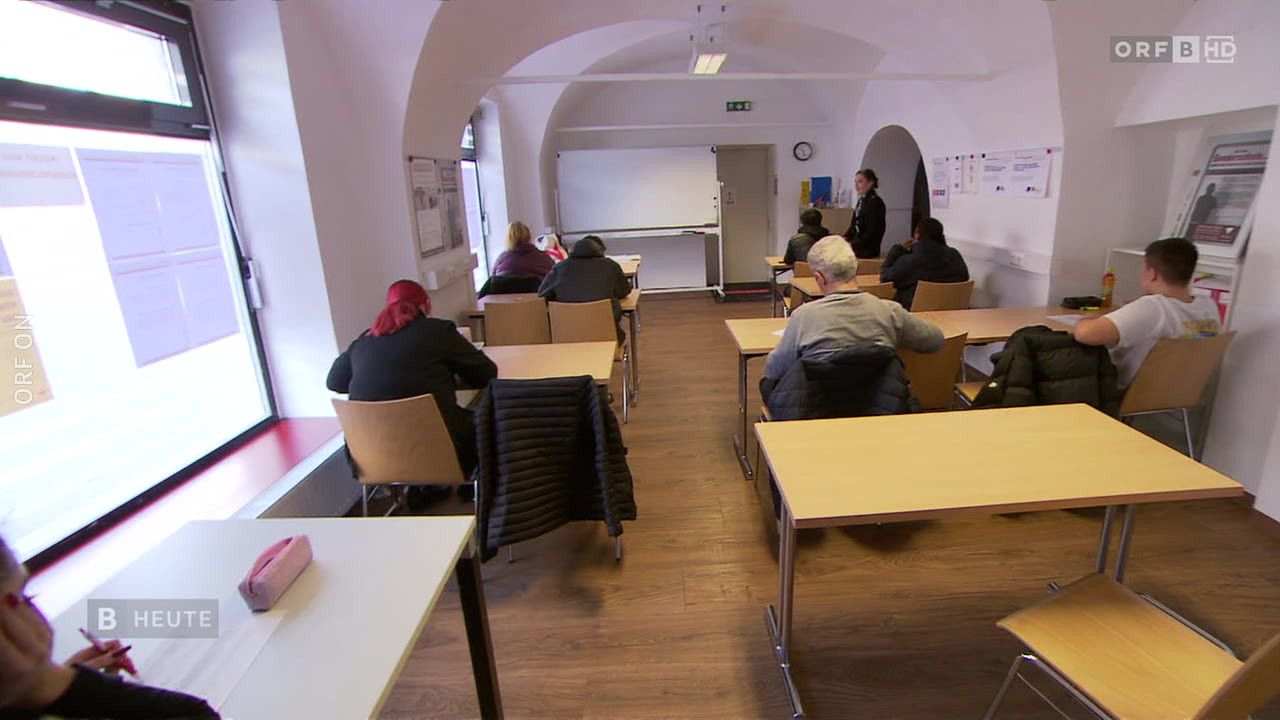 Ein Klassenzimmer mit gewölbten Decken, Schülern in Reihen und einer Lehrerin, die an einer Tafel steht. An den Wänden hängen Zettel.