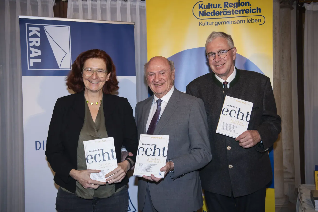 Drei Personen stehen zusammen, lächeln und halten Bücher in den Händen. Die Frau auf der linken Seite trägt eine Brille und eine Halskette. Der Mann in der Mitte trägt eine Krawatte und hat eine Brille auf. Der Mann auf der rechten Seite trägt einen Anzug und eine Brille. Hinter ihnen ist ein gelber Banner mit Text.