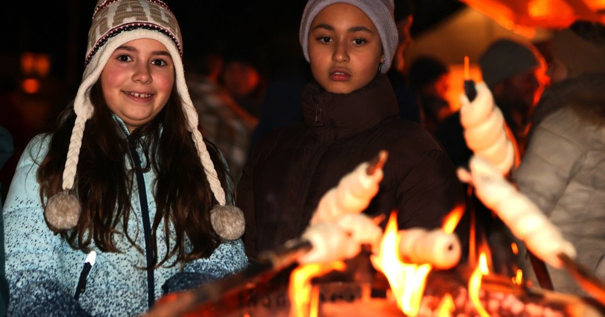 Zwei Mädchen stehen am Lagerfeuer und rösten Marshmallows. Sie tragen Winterkleidung und lächeln.