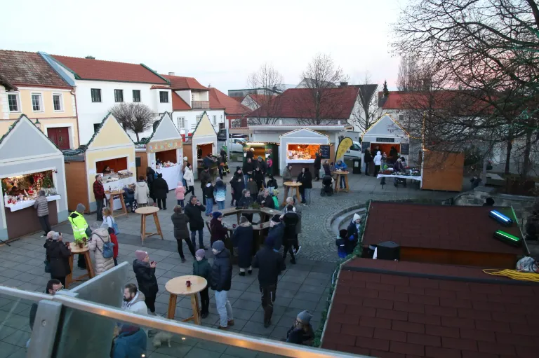 Ein belebter Platz mit festlichen Marktständen, Menschen in Winterkleidung und von Straßenlaternen beleuchtet. Im Hintergrund befinden sich Bäume und mehrere Gebäude.