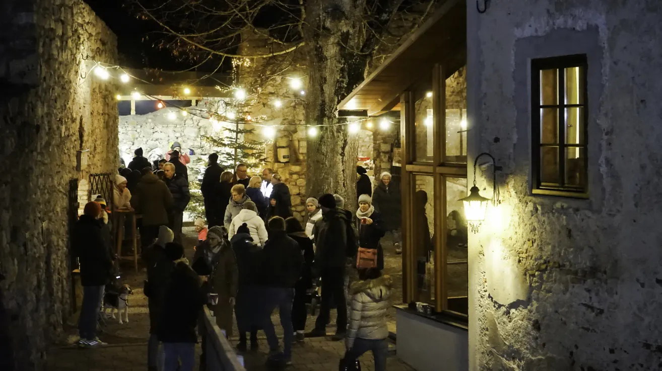 Eine Menschenmenge, warm gekleidet, versammelt sich in einer festlichen Straßenumgebung bei Nacht, geschmückt mit beleuchteten Dekorationen und einem Weihnachtsbaum.