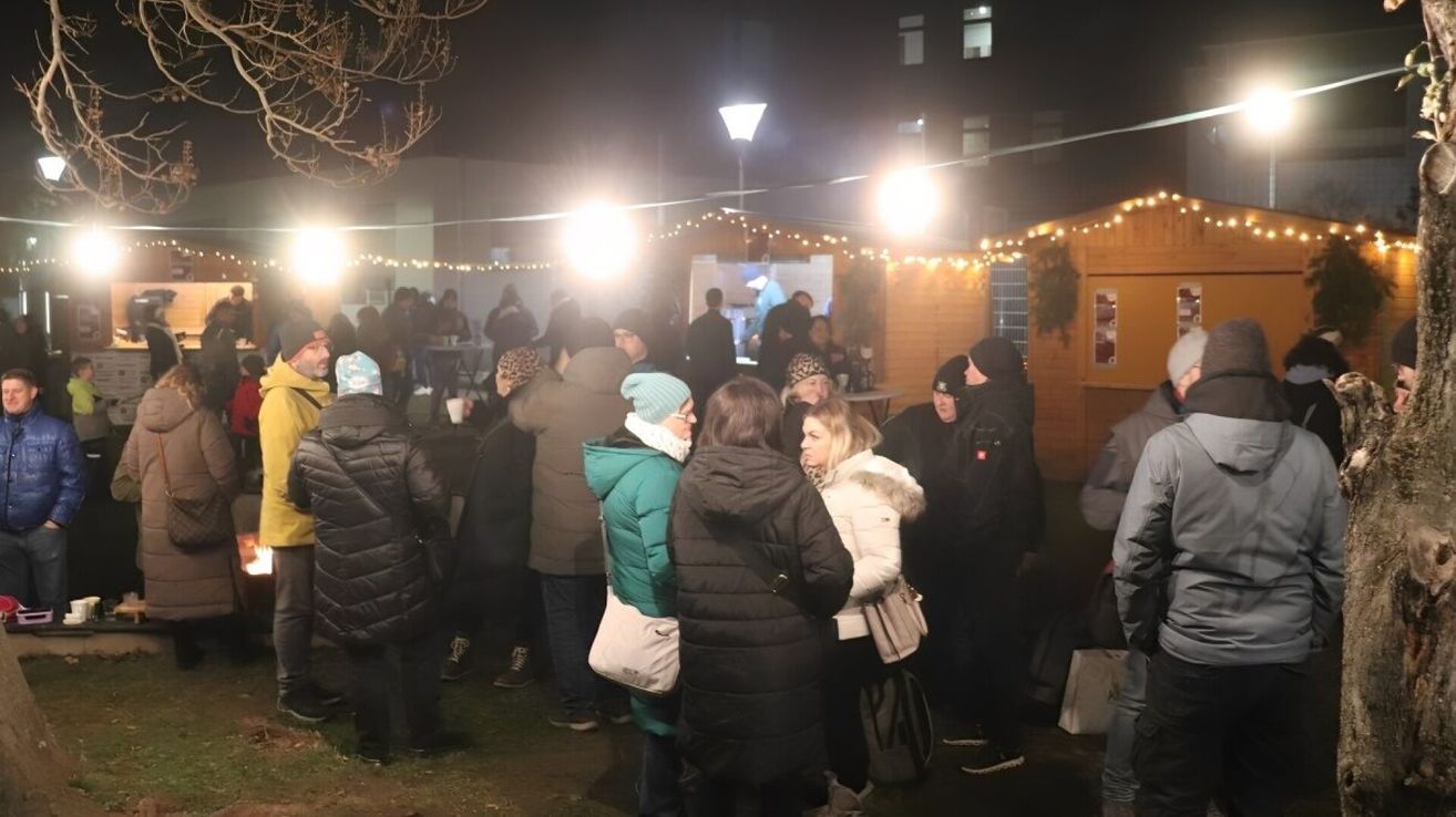 In der Nacht versammeln sich eine Gruppe von Menschen in Winterkleidung vor einem Gebäude, einige mit Tassen, unter dekorativen Lichtern.