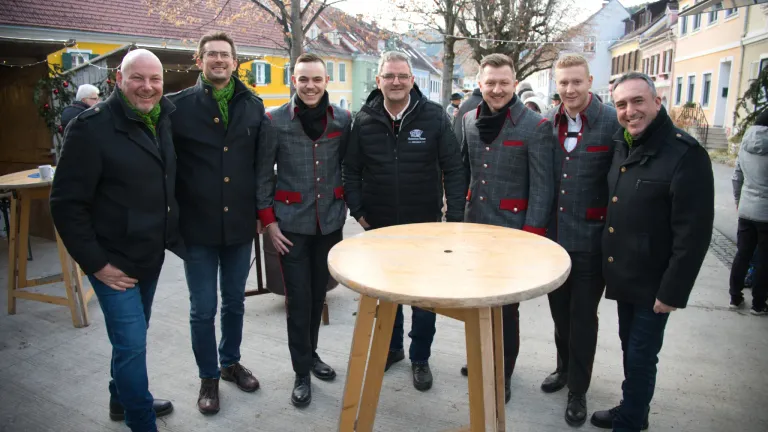 Fünf Männer in traditioneller Kleidung stehen um einen Holztisch in einer Außenumgebung. Der Mann in der Mitte trägt einen schwarzen Mantel. Hinter ihnen stehen Gebäude und Bäume.