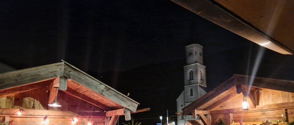 Eine Nachtszene mit einem Kirchturm im Hintergrund und zwei Holzhütten mit Lichtern auf dem Dach.