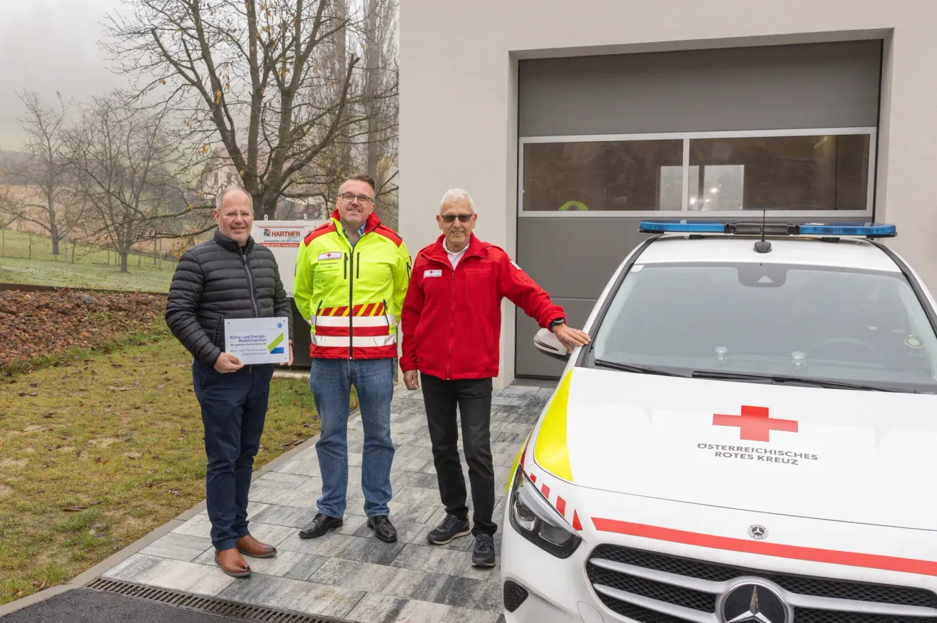 Drei Männer stehen vor einer Garage. Einer hält ein Zertifikat, ein anderer berührt einen roten Kreuz-Ambulanz. Der dritte Mann lächelt.