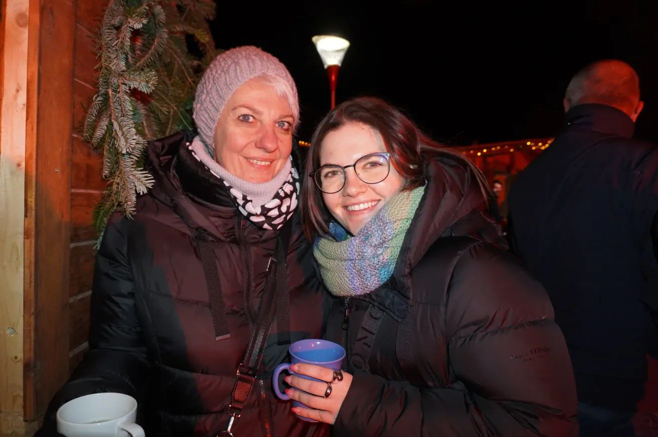 Zwei Frauen posieren nachts draußen für ein Foto, in Wintermänteln und Schals. Eine Frau hält eine blaue Tasse, und ein Weihnachtsbaum ist im Hintergrund zu sehen.