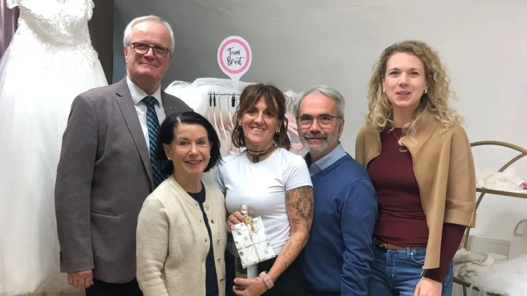 Fünf Personen stehen lächelnd zusammen vor einer Wand. Eine Person hält ein Geschenk. An der Wand befindet sich ein Schild mit der Aufschrift Team Braut.