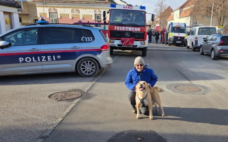 Eine Frau kniet mit ihrem Hund vor einem Polizeiauto. Hinter ihr sind Rettungsfahrzeuge geparkt. Gebäude säumen die Straße.